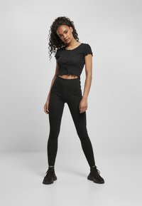 Zwarte cropped t-shirt met korte, gerimpelde mouwen en knoopdetails, gecombineerd met hoge zwarte leggings en zwarte sneakers.