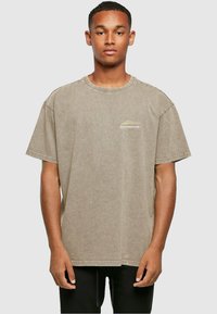 Tricou gri oversized cu mânecă scurtă din material bumbac, cu guler rotund și un mic logo pe partea stângă a pieptului. Textură moale.