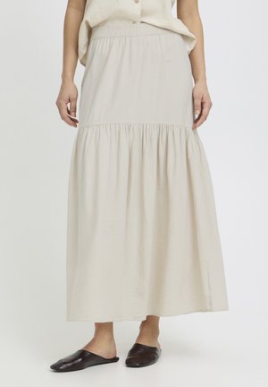 Kobieta ubrana w długą, jasno-beżową marszczoną spódnicę z elastycznym paskiem i czarne wsuwane baleriny, stojąca na białym tle.