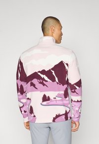 Pullover in pile rosa con grafica di montagne viola e paesaggio. Caratterizzato da un colletto alto e maniche lunghe con orli a polsino. Texture morbida.