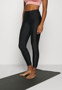 Kvinna i svarta högmidjade leggings och en rosa sportbh, stående barfota på en svart yogamatta mot en vit bakgrund.