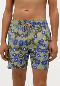 Shorts de bain verts avec des motifs géométriques bleus et navy. Taille élastique avec un cordon de serrage beige. Longueur atteignant le mi-cuisse.