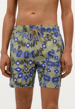 Shorts de bain verts avec des motifs géométriques bleus et navy. Taille élastique avec un cordon de serrage beige. Longueur atteignant le mi-cuisse.