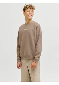 Sudadera marrón con cuello redondo y mangas largas, que presenta un pequeño logo blanco en el pecho y un parche texturizado en la manga.
