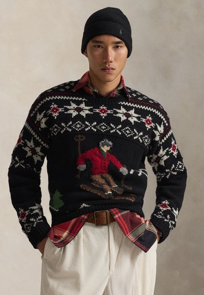 Pull en tricot noir avec des motifs blancs, rouges et verts. Présente un motif de skieur en rouge et gris. Superposé avec une chemise à carreaux et un pantalon beige.