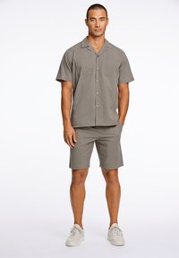 Grå kortärmad skjorta med knappar och två bröstfickor, i kombination med matchande shorts. Modellen bär vita sneakers. Enkelt texturerad tyg.