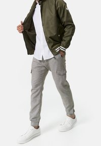 Olijfgroene bomberjack met witte strepen, witte overhemden, grijze joggingsbroek met zijzakken en witte sneakers.