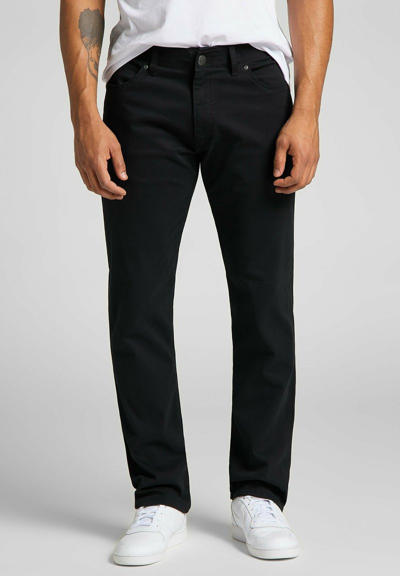 Jeans de mezcla de algodón negros con corte recto, diseño de cinco bolsillos, presillas para cinturón y un acabado sutil. Combinados con zapatillas blancas.