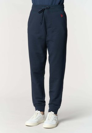 Pantaloni jogger blu navy con vita regolabile a coulisse, tasche laterali, logo ricamato rosso, indossati con sneakers bianche, mostrati dalla vita ai piedi.