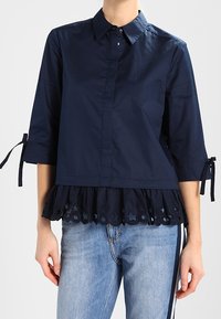 Marinblus med krage, trekvartslånga ärmar och volangkant med utskurna stjärnmönster. Slät tygtextur.