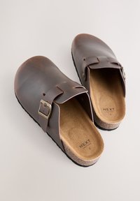 Braune Leder-Clogs mit abgerundetem Zeh, verstellbarem Riemen und weichem braunem Innenfutter. Korksohle für Dämpfung und Gummilaufsohle für Griffigkeit.
