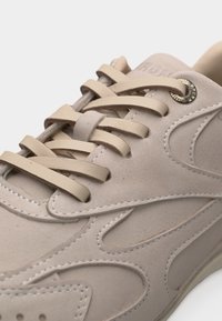 Zbliżenie beżowego trampka z grubymi płaskimi sznurowadłami, wykonany z zamszu, z precyzyjnymi przeszyciami wokół przelotek i bocznych paneli.