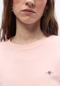 Κοντινό πλάνο μιας γυναίκας που φοράει ανοιχτό ροζ φούτερ με ένα μικρό λογότυπο GANT στο στήθος και ασημένια σκουλαρίκια κρίκους.