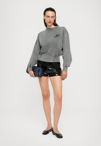 Felpa grigia con una vestibilità cropped e logo ricamato, abbinata a pantaloni corti neri in vernice. Il modello tiene una pochette blu texturizzata e indossa ballerine nere.