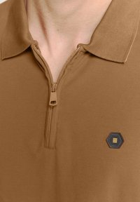 Bruine rits polo shirt van textuurstof, met een hexagonaal insigne met zwarte en gele accenten op de linkerborst.