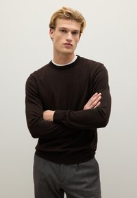 Maglione marrone a maglia con colletto rotondo, maniche lunghe e polsi a costine, indossato sopra una camicia bianca, abbinato a pantaloni grigi.