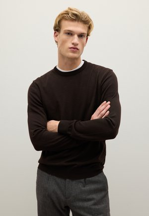 Maglione marrone a maglia con colletto rotondo, maniche lunghe e polsi a costine, indossato sopra una camicia bianca, abbinato a pantaloni grigi.