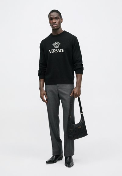 Černý pletený svetr s logem Versace, šedé kalhoty na míru, černé kožené boty a černá polstrovaná kabelka přes rameno se zlatými detaily.