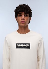 Beige t-shirt met lange mouwen, gemaakt van katoen, met een zwart rechthoekig logo waarop "NAPAPIJRI" en "geographic" staat. Soepele textuur.
