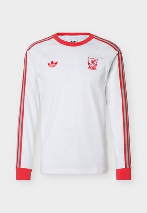 Bijela sportska majica s dugim rukavima s crvenim obrubom, tri crvene pruge na rukavima, crveni Adidas logo na lijevoj strani prsa i grb Liverpool FC na desnoj strani prsa.