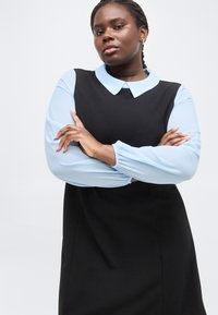 Αυτοπεποίθητη γυναίκα με πλεξούδες, φορούσα μαύρο φόρεμα με ανοιχτό μπλε μανίκια και γιακά, με σταυρωμένα τα χέρια, σε απλό λευκό φόντο.