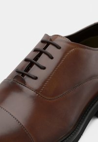 Chaussure habillée en cuir marron à finition lisse, dotée d'un bout arrondi, d'un design à lacets et d'une semelle noire contrastante avec des détails de couture.
