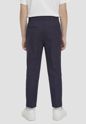 Persona che indossa pantaloni blu navy su misura con tasche posteriori, calzini bianchi, scarpe da ginnastica bianche e camicia bianca a maniche corte, in piedi di spalle.