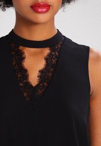 Haut noir avec un col montant, insert en dentelle en V et sans manches, fabriqué dans un tissu lisse avec un léger brillant.