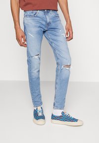 Man som bär ljusblå slitna jeans och blå mönstrade sneakers med vita sulor och snörning, stående mot en neutral bakgrund.