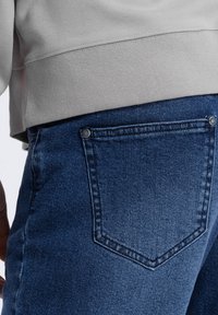 Tummansiniset farkut, joissa on kevyt stretch, ja joissa on takatasku, jossa on sävysuunnittelua ja metallinitteitä. Asusteena vaaleanharmaa neule.