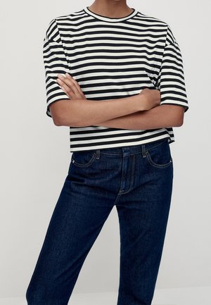 Osoba ubrana w czarno-białą koszulkę w poziome paski z krótkim rękawem, z założonymi rękami i ciemnoniebieskie dżinsy na jednolitym tle.