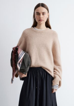 Femme aux longs cheveux bruns portant un pull beige en tissu pelucheux sur une chemise rayée, tenant un sac gris foncé avec des foulards à motifs, et une jupe noire.