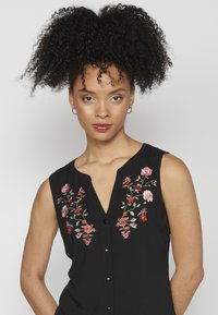 Blusa nera senza maniche con ricami floreali in rosso e rosa, con scollatura a V e chiusura frontale a bottoni. Tessuto dalla texture liscia.