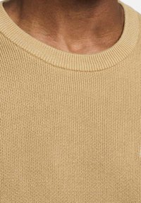 Närbild av hals och övre bröst som bär en beige stickad tröja med rundad hals, som visar textur och detaljer med ribbat halsband.