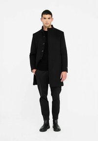 Manteau en laine noire à coupe droite, col montant et fermeture à boutons. Associé à un pull texturé noir et un pantalon taille ajustée. Bottines noires.