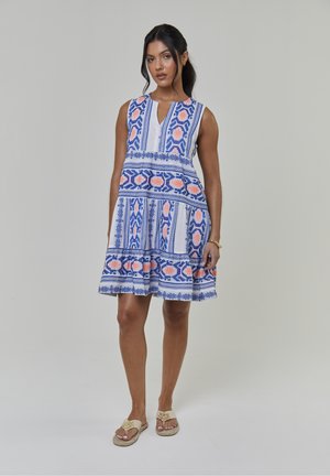 Robe blanche sans manches avec motifs floraux bleus et corail, ourlet évasé et tissu texturé. Portée avec des sandales plates marron clair.