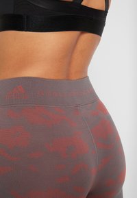 High-waisted leggings in a gray-brown color with a red camouflage pattern and a textured waistband featuring the Adidas and Stella McCartney logos.