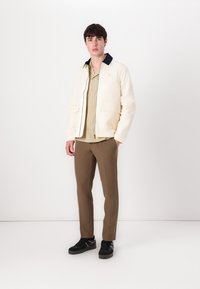 Cream canvas jacket with navy collar, paired with a beige button-up shirt and brown trousers. Black sneakers complete the outfit.