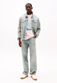 Veste en jean bleu clair avec poches sur la poitrine, portée sur un t-shirt graphique rose représentant une scène de plage colorée, assortie à un jean clair et à des baskets blanches.