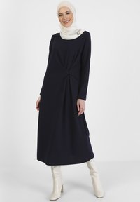 Navy long-sleeve dress with gathered waist detail, paired with a light-colored hijab and cream heeled boots. Smooth fabric texture.