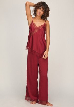 Vrouw met krullend haar, die een rood kanten mouwloos topje en wijde broek draagt, staat blootsvoets tegen een effen achtergrond.