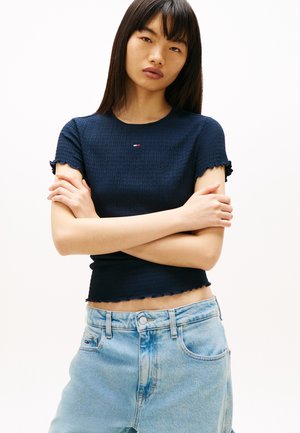 Ung kvinde med glat sort hår iført en tætsiddende mørkeblå top med korte ærmer og lyseblå højtaljede jeans, med armene krydset.