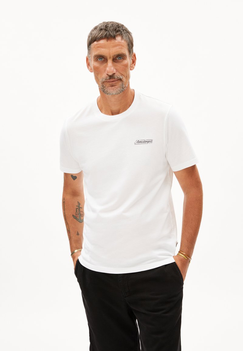 Weißes Baumwoll-T-Shirt mit Rundhalsausschnitt, kurzen Ärmeln und einem kleinen aufgedruckten Logo auf der linken Brust. Getragen mit schwarzen Hosen.