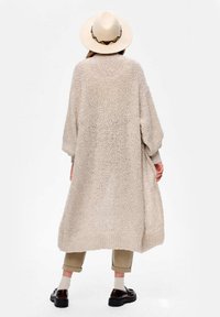 Długi, oversize'owy beżowy sweter kardigan o fakturowanym wykończeniu, z opuszczonymi ramionami i luźną sylwetką. Połączony z ciemnymi butami i spodniami.