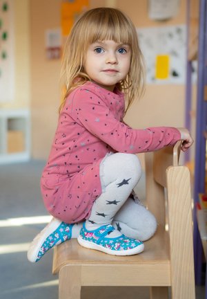 Kind in einem pinken Kleid mit Sternenmuster, grauen, mit Sternen gemusterten Strumpfhosen und blauen Schuhen mit Cartoon-Motiven, sitzt auf einem Holzstuhl.