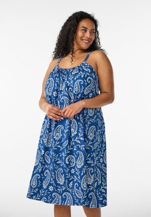 Smilende kvinde med krøllet hår iført en ærmeløs blå kjole med hvid paisley- og blomsterprint, står foran en ensfarvet hvid baggrund.