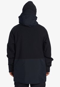 Zwarte lange jas met een capuchon, voorzien van een gestructureerd bovenstuk en een glad onderstuk. Draagt een donkere gebreide beanie.