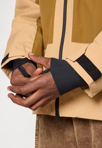 Χέρια που ρυθμίζουν μαύρα μανίκια με άνοιγμα για τον αντίχειρα κάτω από μια μπεζ ζακέτα με μαύρο φερμουάρ και βέλκρο μανσέτες πάνω από καφέ παντελόνι.