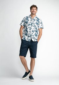 Chemise à manches courtes avec un motif de palmier blanc et bleu, associée à un short marine et des chaussures à enfiler sombres. Fond uni.