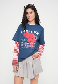 Μπλε γραφική κοντομάνικη μπλούζα με κόκκινο λουλουδάτο σχέδιο, λευκό κείμενο και μακριά μανίκια με κόκκινες ρίγες· συνδυασμένη με ανοιχτό γκρι πλισέ φούστα.